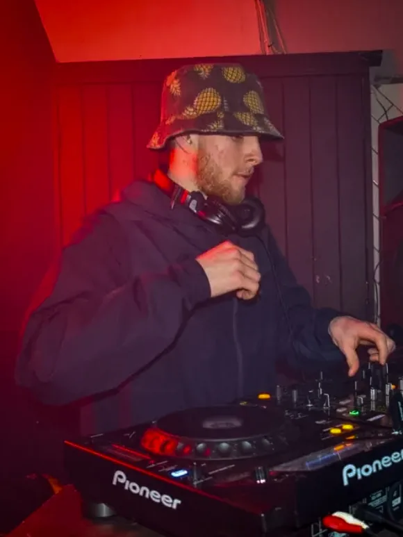 NATUS wearing a patterned bucket hat and headphones while mixing under red lighting