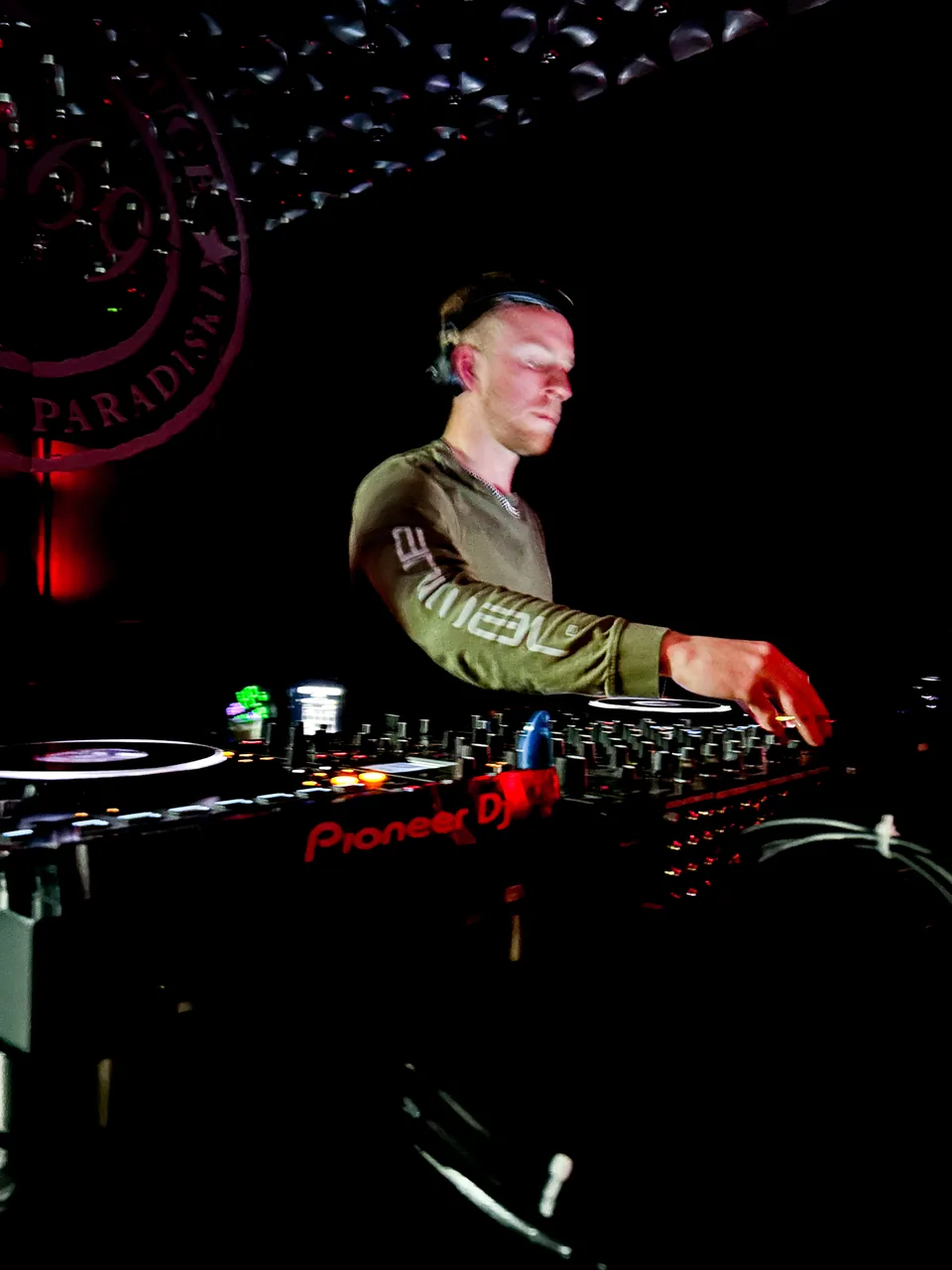 NATUS wearing headphones and adjusting Pioneer DJ decks in a dark venue